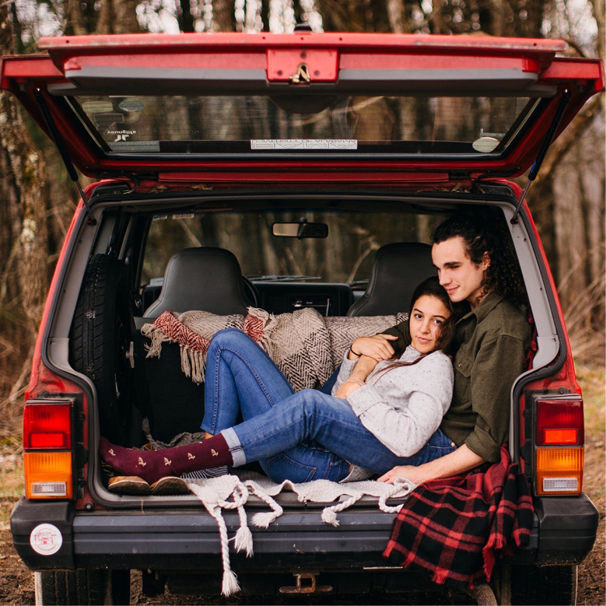 A couple snuggle together in blankets in the back of a red Jeep Cherokee.