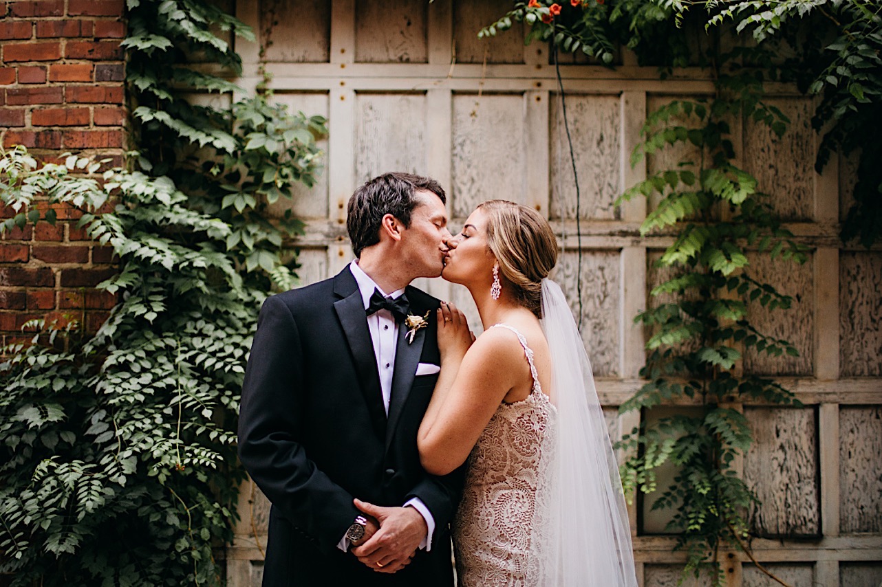 bride and groom kiss in front of ivy-covered white wooden wall