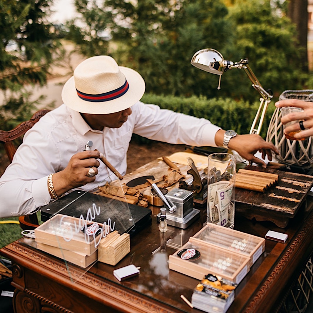 A cigar maker rolls a cigar at a table on a landscaped lawn. 