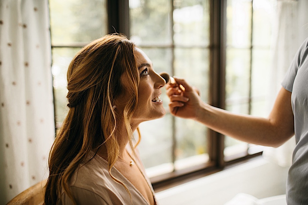 A bride smiles as her makeup is touched up on her wedding day.