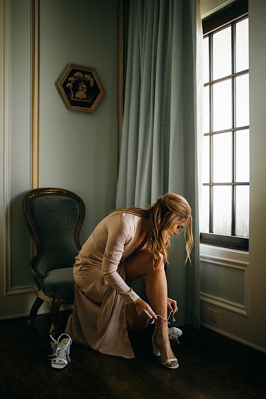 A bride sits on a vintage chair next to a window and fastens her shoes.