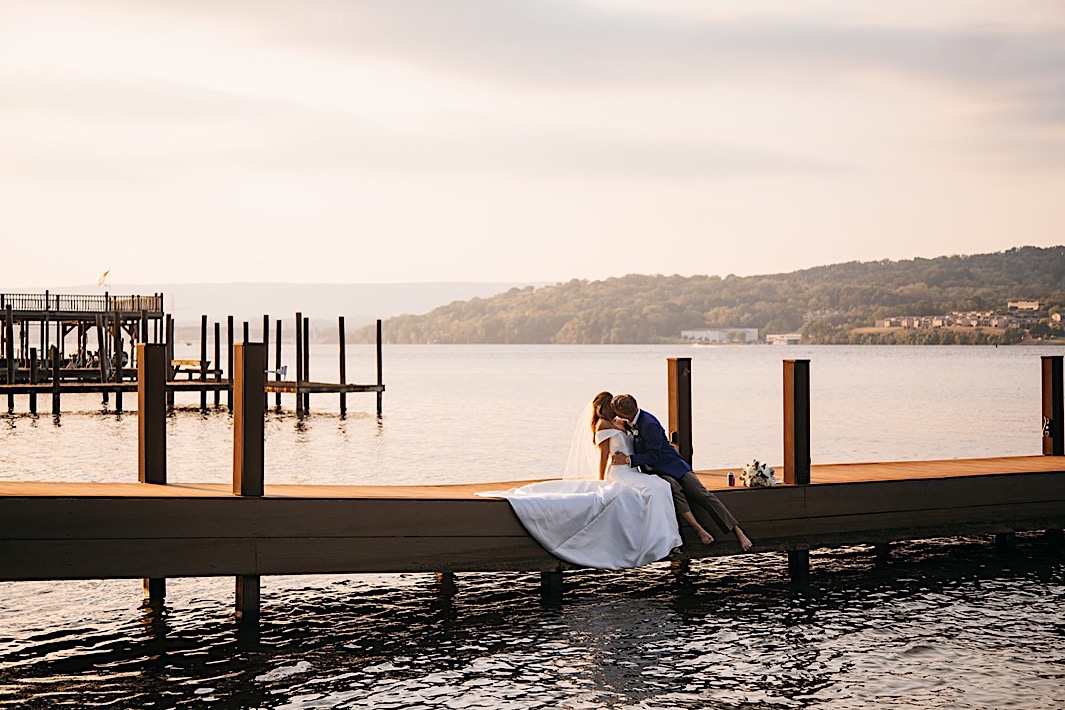 A bride and groom kiss on the edge of a dock.