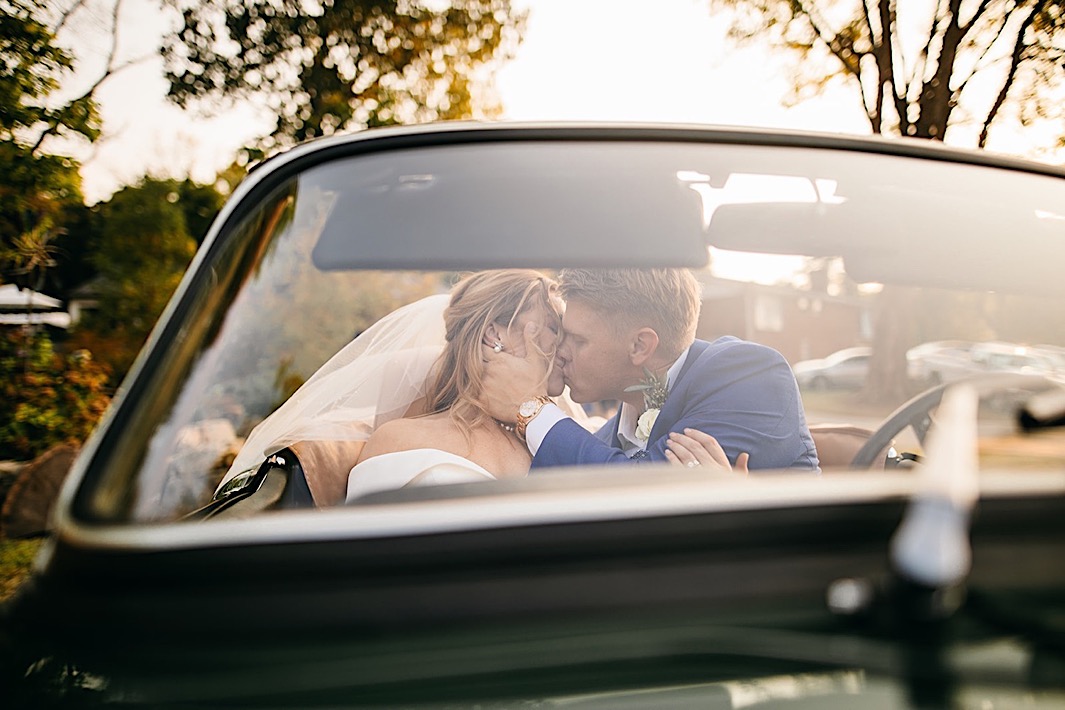 A bride and groom kiss in the front seat of a vintage car.