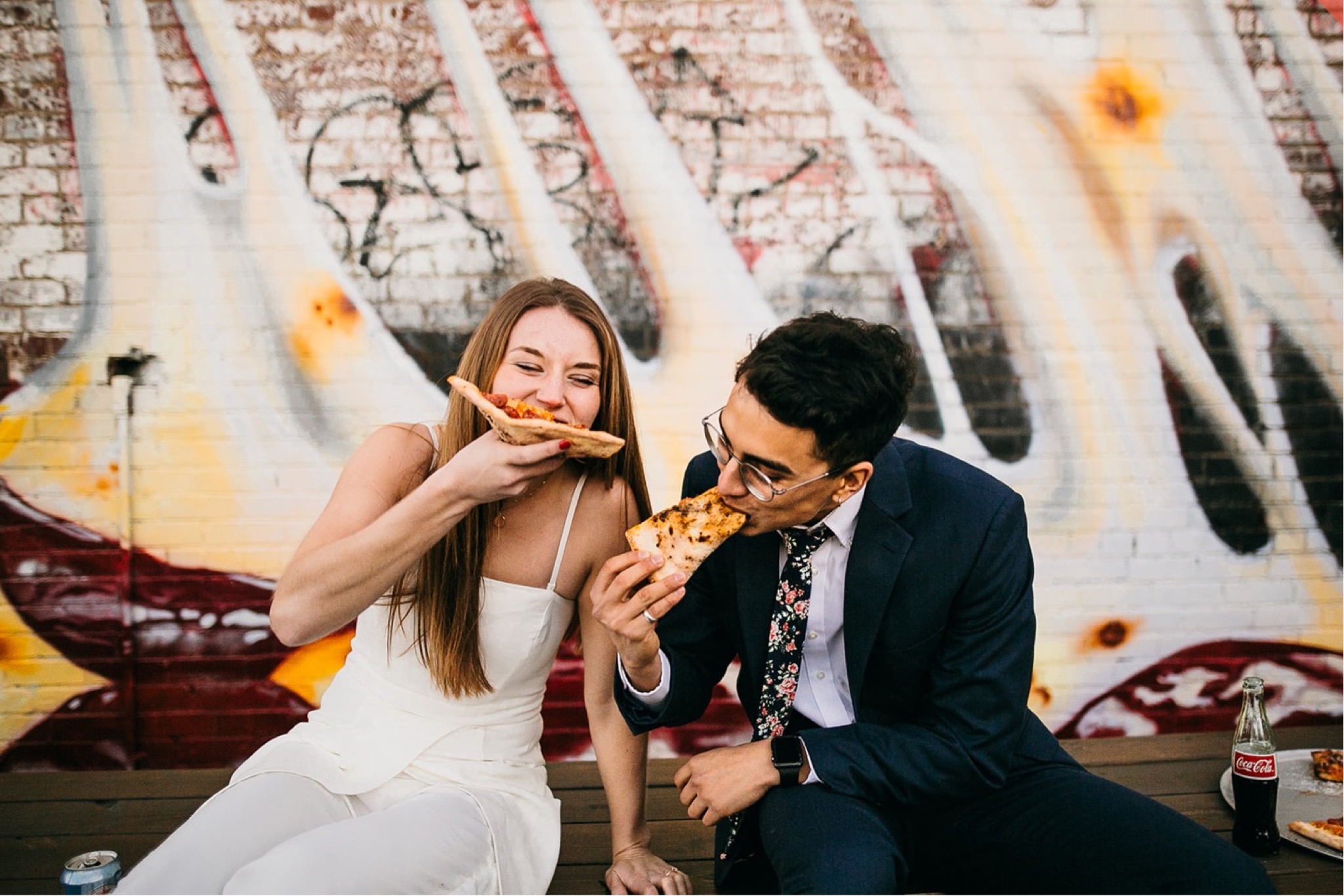 Man and woman share pizza in front of a massive pizza mural.