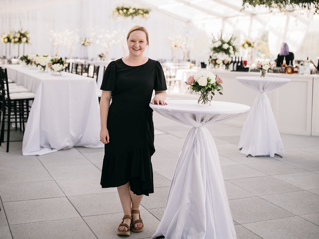 A wedding planner smiles and leans against a cocktail table in an empty reception space.