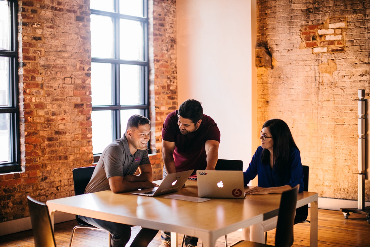 Three coworkers huddle around laptops in a bright office full of exposed brick.