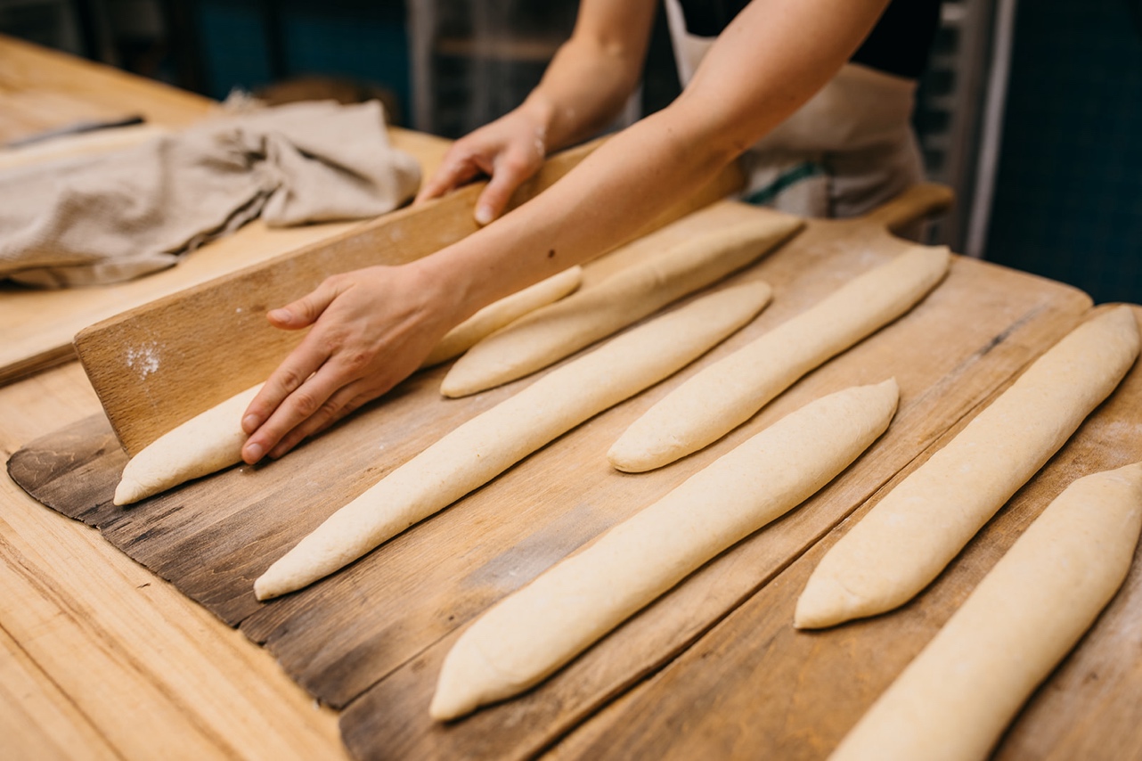 A baker forms long, thin loaves of bread on a wooden surface.