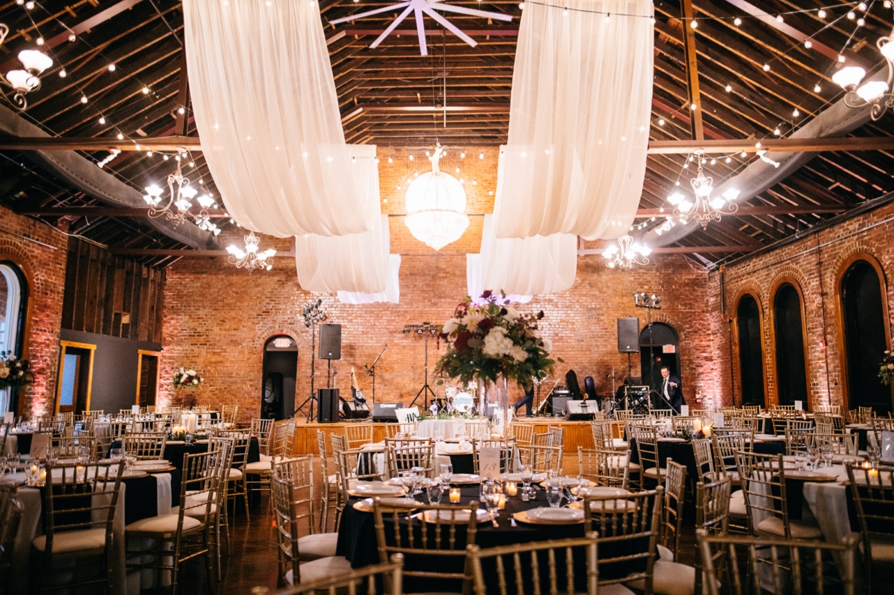 A brick reception venue set for a wedding reception with white drapes hanging from the ceiling.