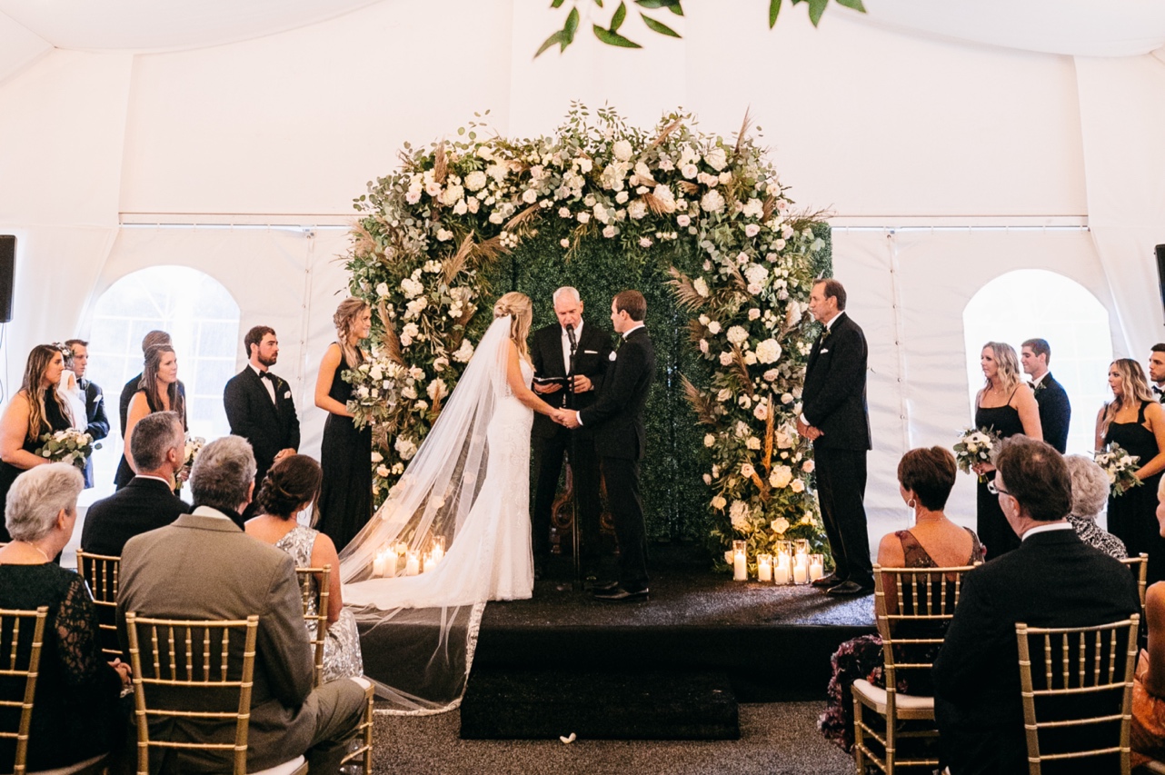 Bride and groom hold hands at the altar beneath an arbor of white and green florals.