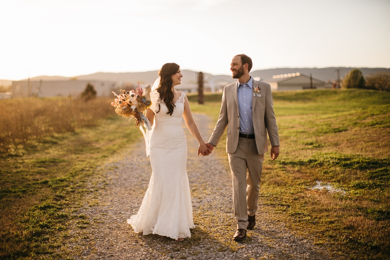 A bride holding a bouquet of muted, autumn-toned florals as she walks through a field with her groom.
