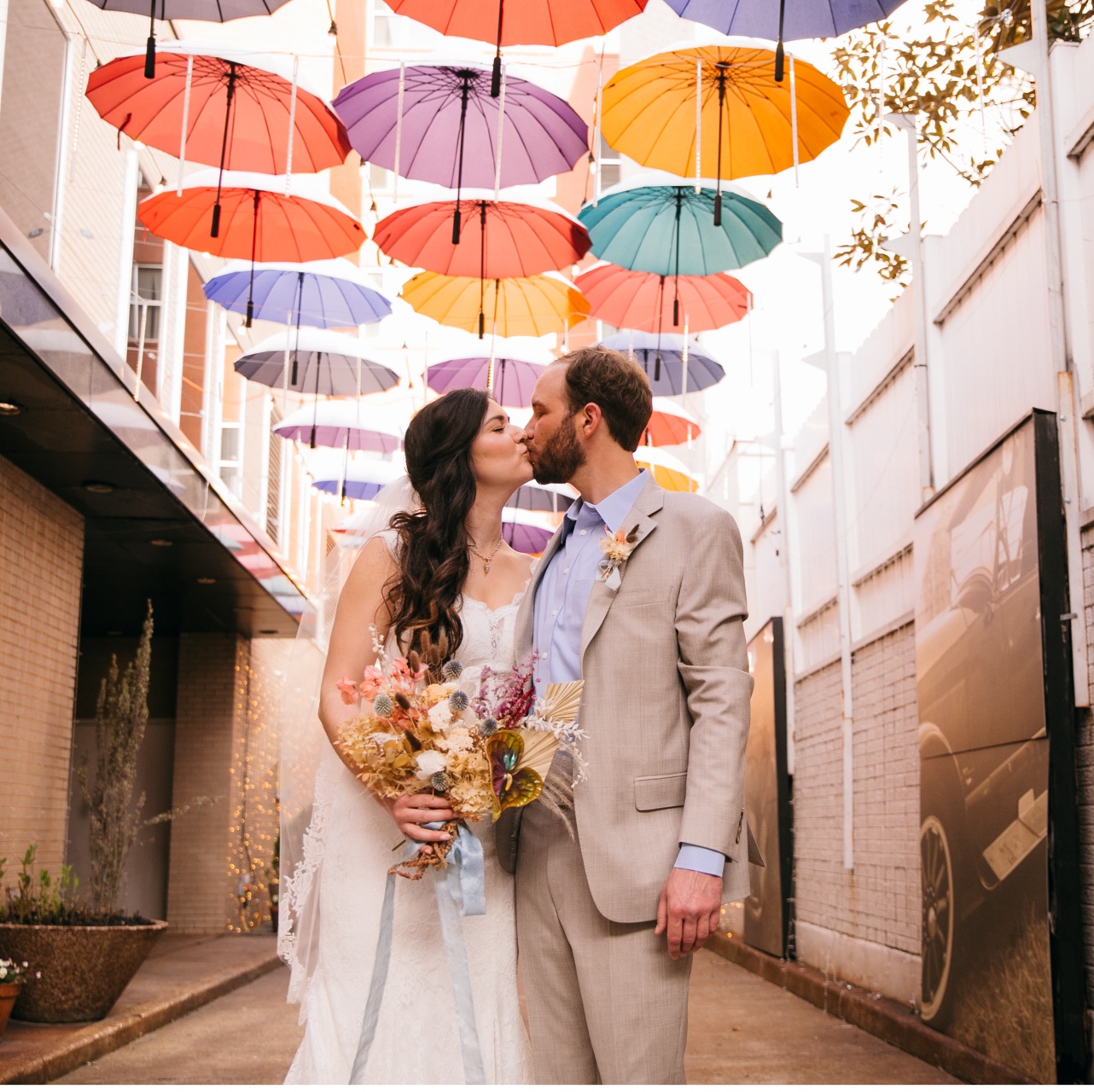 Bride and groom kiss in an alley under an umbrella canopy.