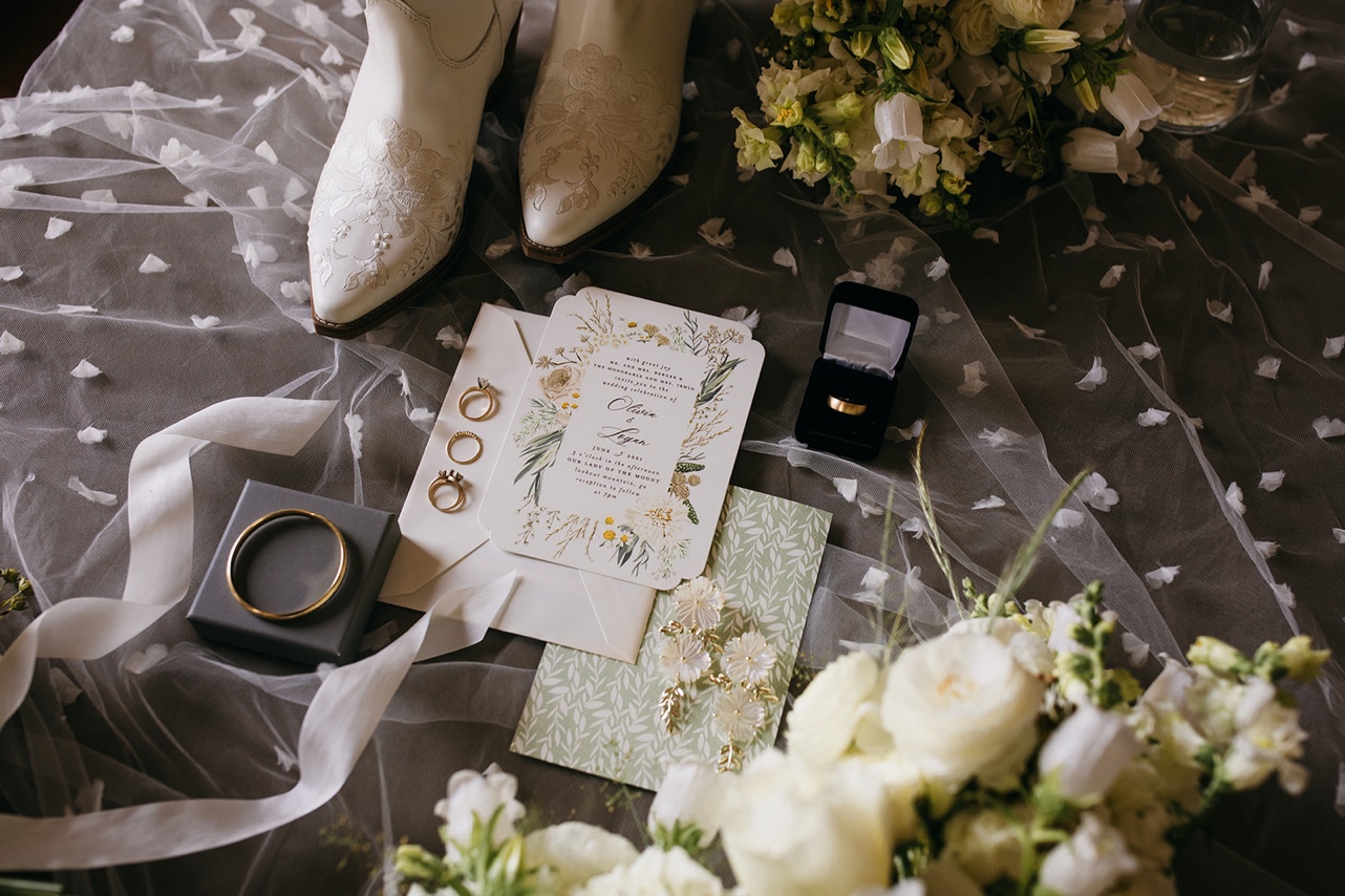 A flatlay of the bride's boots, accessories, and wedding invitations on top of her unique wedding veil.