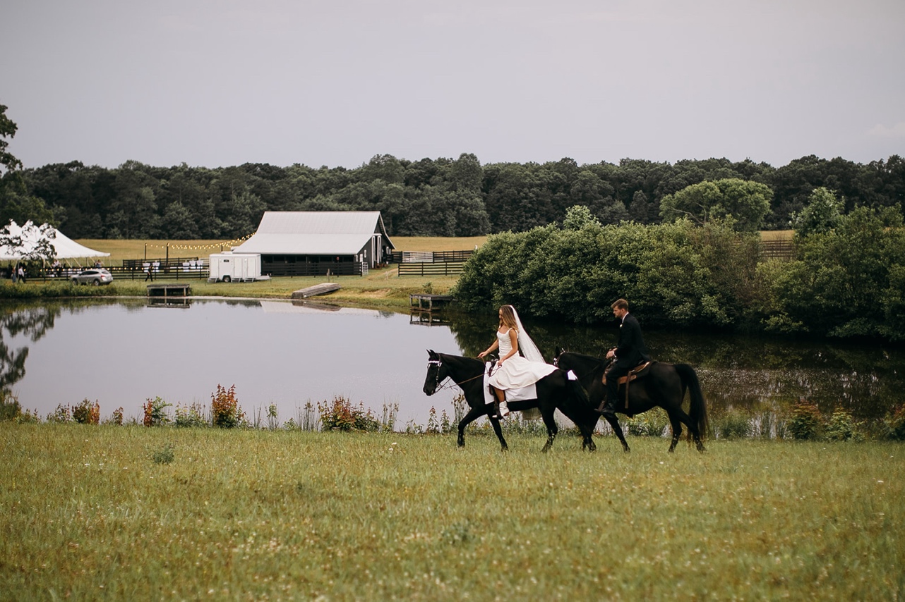 Bride and groom ride black horses side-by-side across a field next to a pond.
