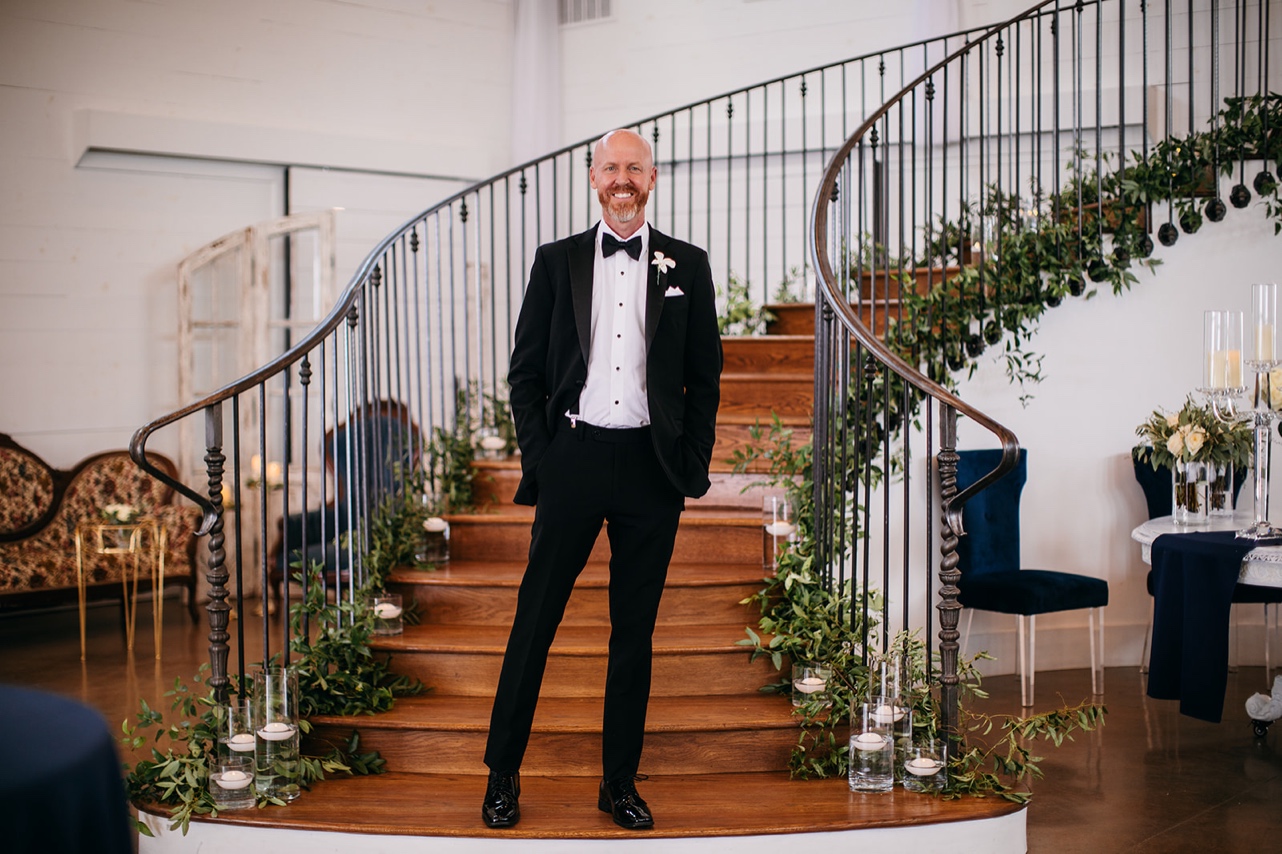 At his Howe Farms wedding, groom stands on a staircase lined with greenery and candles.