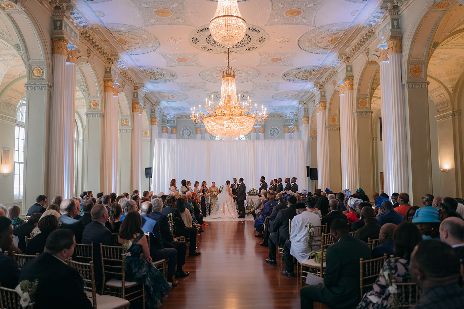 Wide view of the ceremony inside The Biltmore Ballrooms Atlanta with guests gathered beneath crystal chandeliers. 