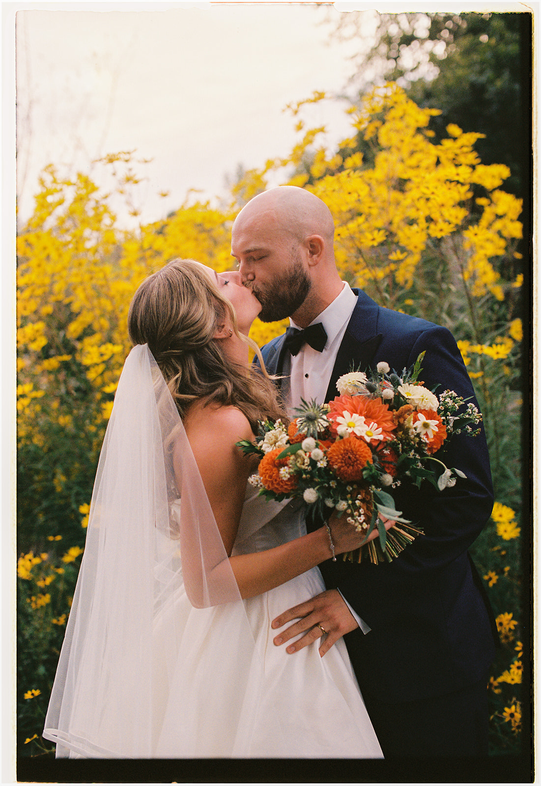 Bride and groom sharing a kiss among vibrant flowers during an NC Arboretum wedding. 